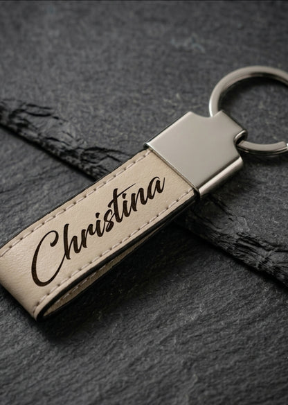 A personalized leather keychain with the name 'Christina' engraved in black script on a beige leather strap, attached to a silver metal cap and split ring, resting on a dark slate background.