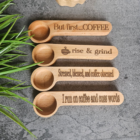 A set of four wooden coffee-themed scoops and clips, with text on them, placed on a gray surface with greenery in the background.