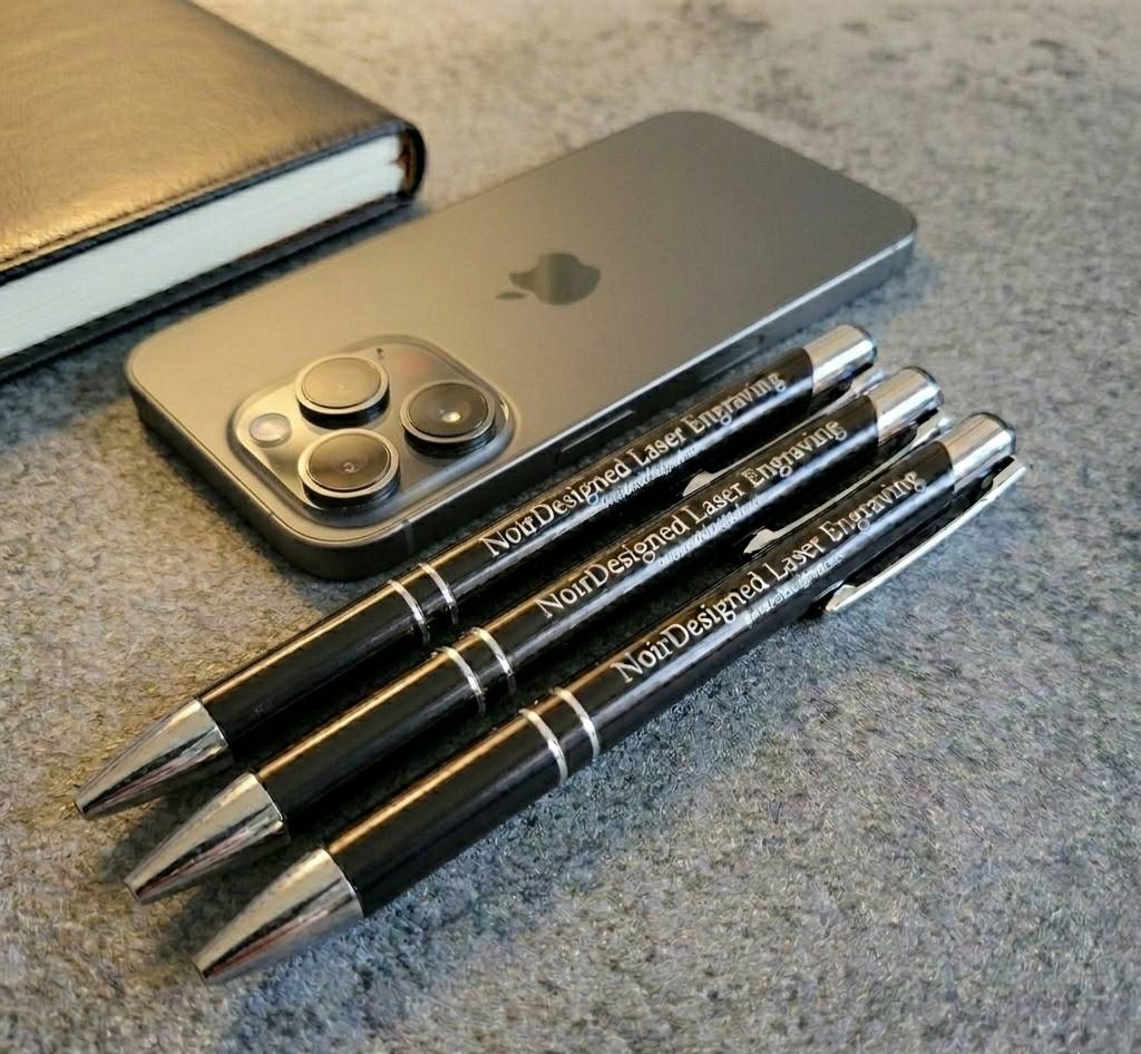 Three black pens with laser engraving on a gray surface next to a smartphone.