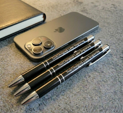 Three black pens with laser engraving on a gray surface next to a smartphone.